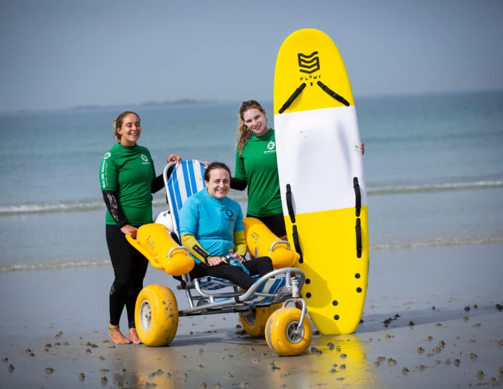Surfers with adaptive beach wheelchair and surfboard on sand.